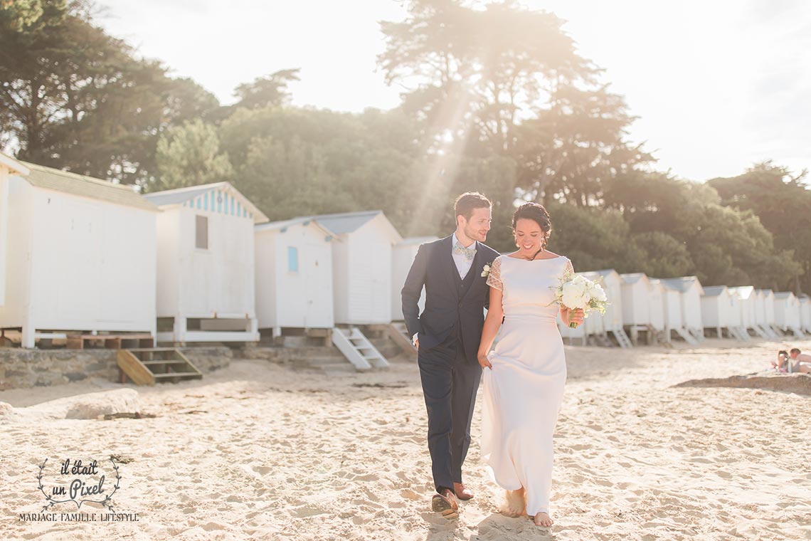 Photo en contre jour d'un couple de mariés marchant sur le plage devant des cabines blanches le jour de leur mariage en Vendée (Ile de Noirmoutier)