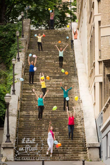 Photo de groupe lors d'un shooting EVJF sur les rives de Seine à Paris avec les participantes les bras en l'air avec des balons colorés et tenues colorées dans de très hauts escaliers