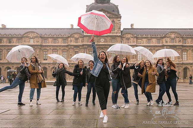 Photo de groupe lors d'un shooting EVJF à dans la cour du Louvre à Paris avec les participantes sous la pluie avec des parapluies transparents
