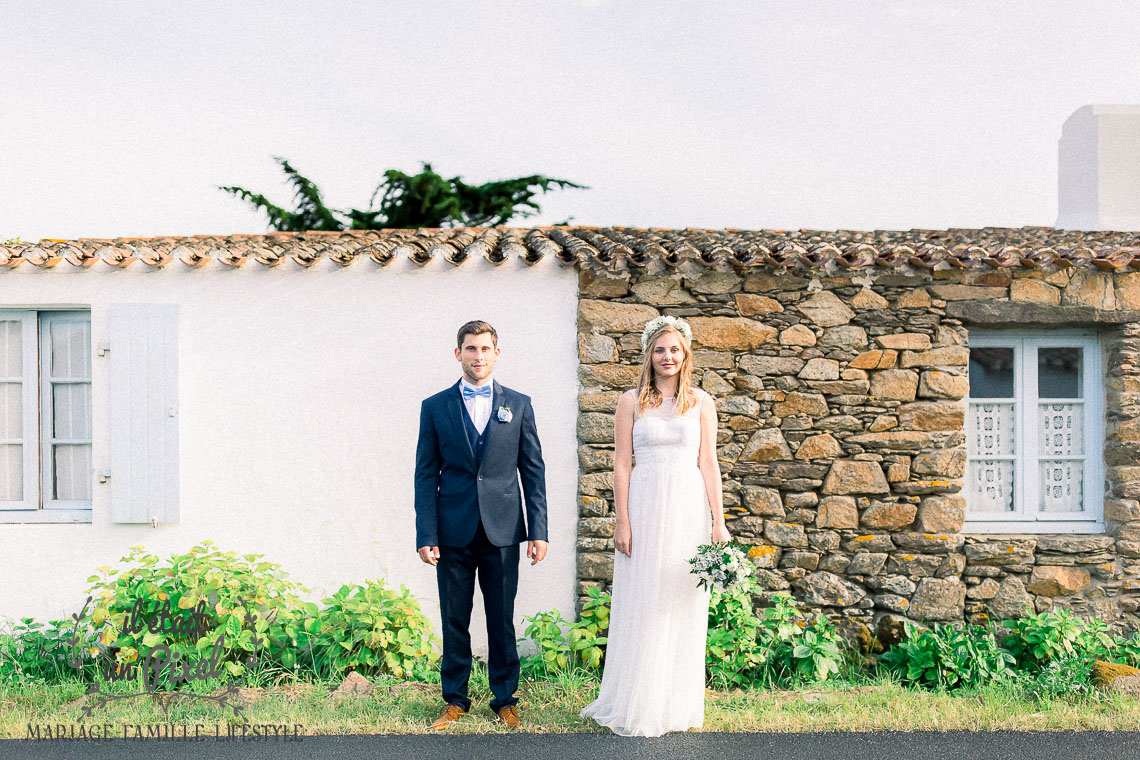 Couple de mariés cote à cote en pose hipster devant un mur blanc pour le marié et foncé pour la mariée