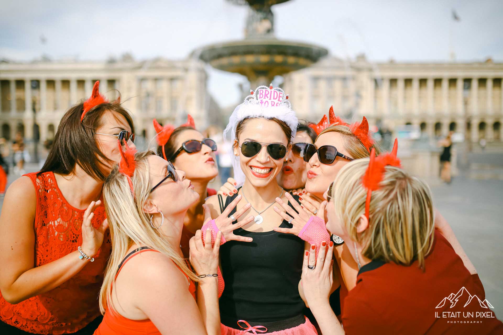 Photo de groupe lors d'un shooting EVJF à la Concorde à Paris avec les participantes en rouge avec oreilles de diable et lunettes de soleil embrassants la future mariée